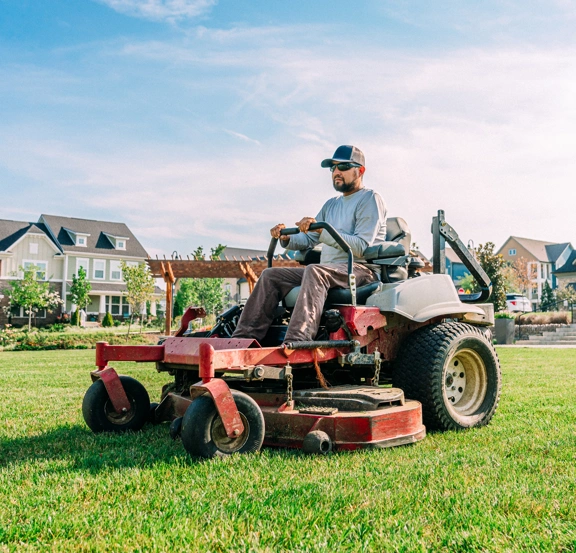 Man driving riding lawnmower, cutting grass in a neighborhood.
